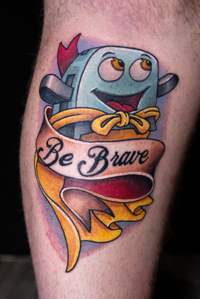 Bright tattoo of cartoon brave little toaster. slightly blue with a yellow cape and a traditional banner that reads "be brave"